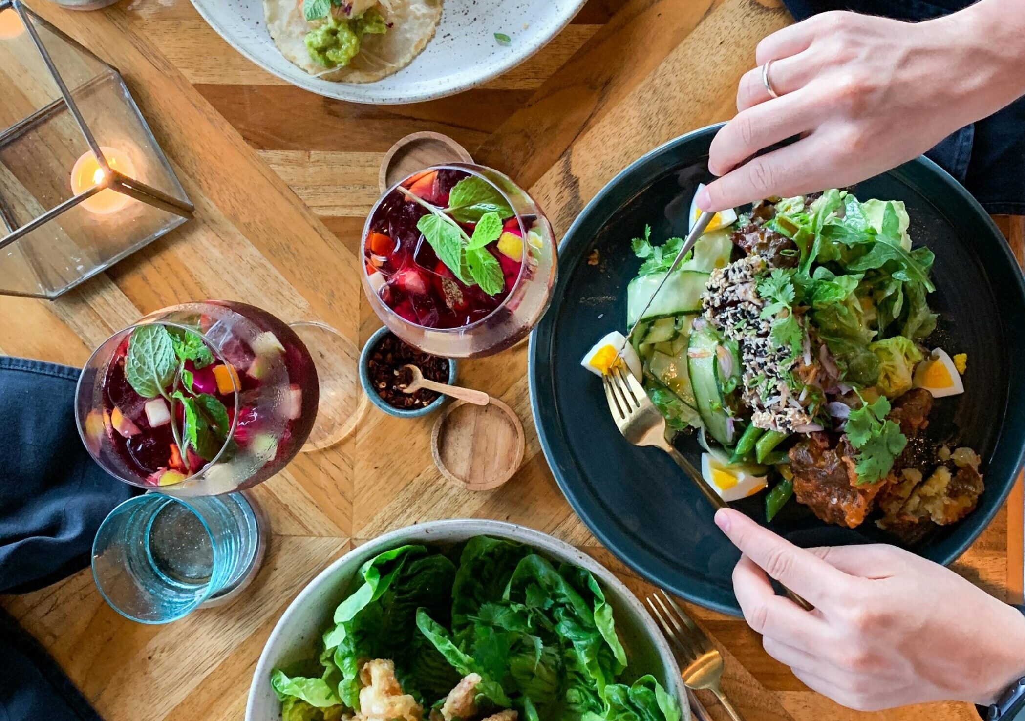 A woman's hands holding a knife and fork over a bowl of fresh salad, with glasses of fruit and additional vegetables surrounding the table.