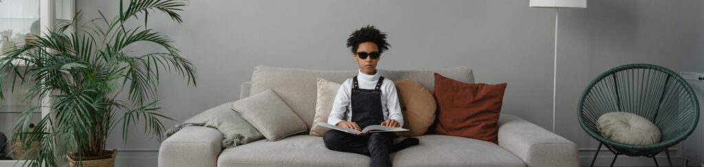 A blind Black girl reading a book in braille while sitting on a couch.